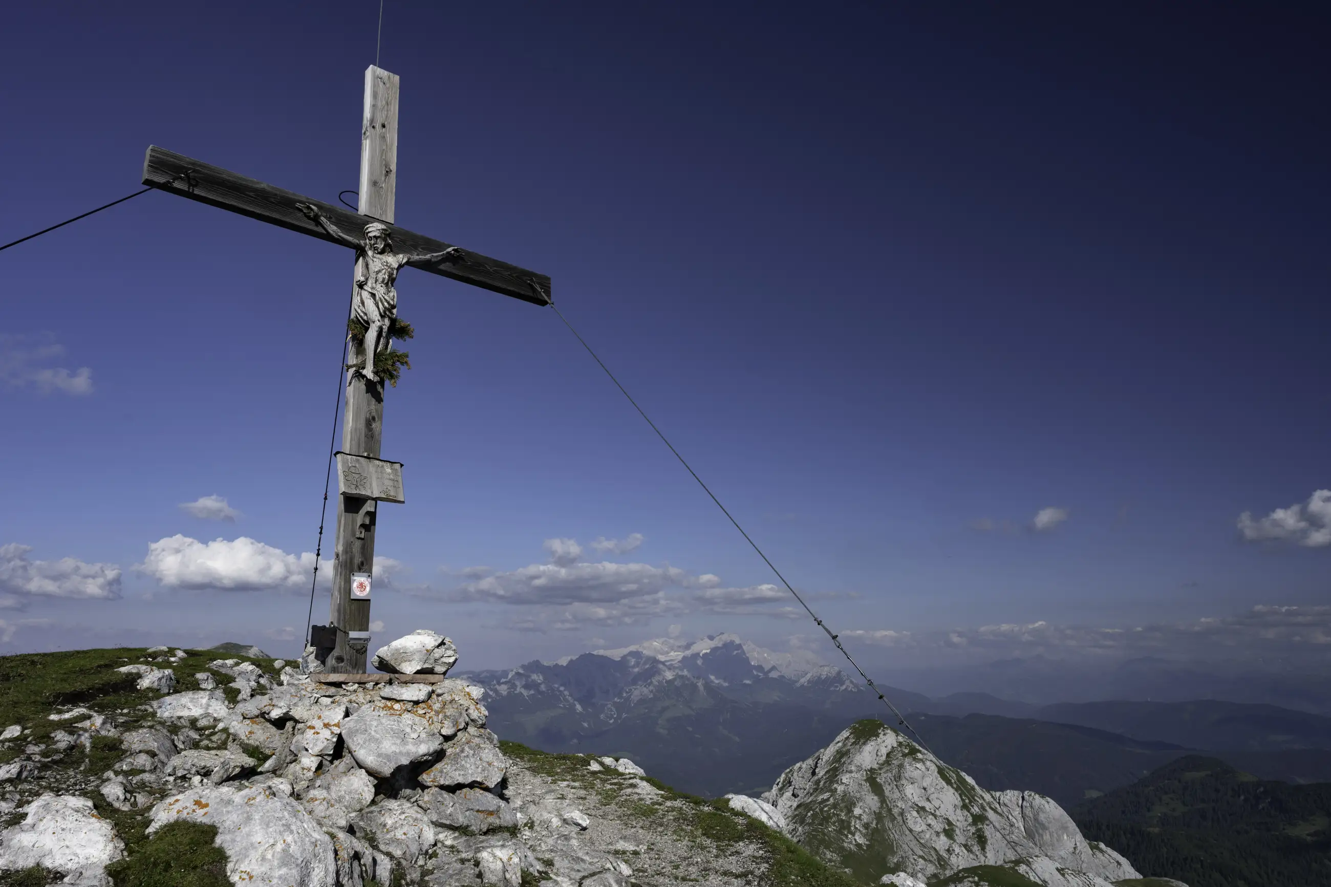 A large wooden cross stands on a rocky mountain summit with panoramic views of distant peaks under a clear blue sky.