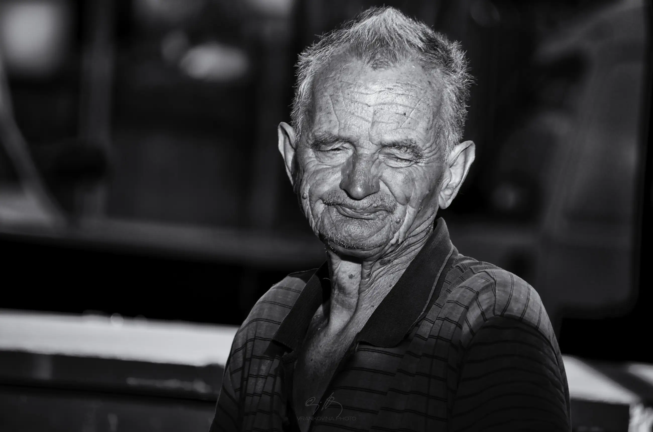 An elderly man with short, gray hair and deep facial wrinkles stands outdoors, wearing a striped collared shirt. He has his eyes closed and a slight, gentle smile on his face. The image is in black and white.