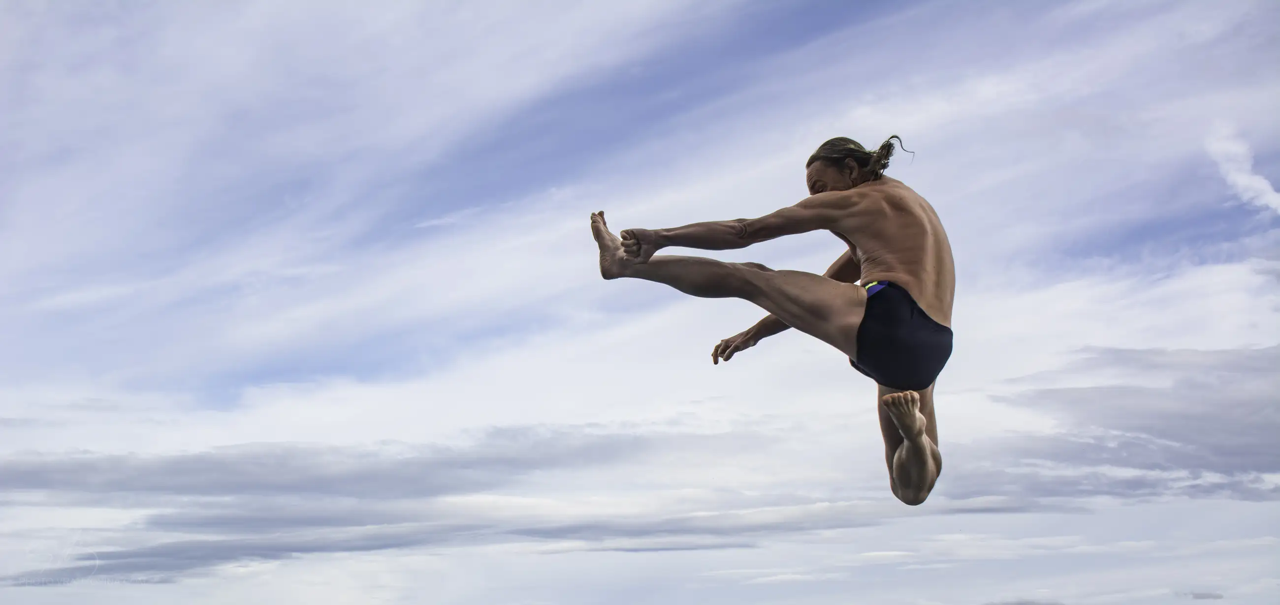 A person wearing swim trunks performs a mid-air split kick against a backdrop of blue sky and clouds.