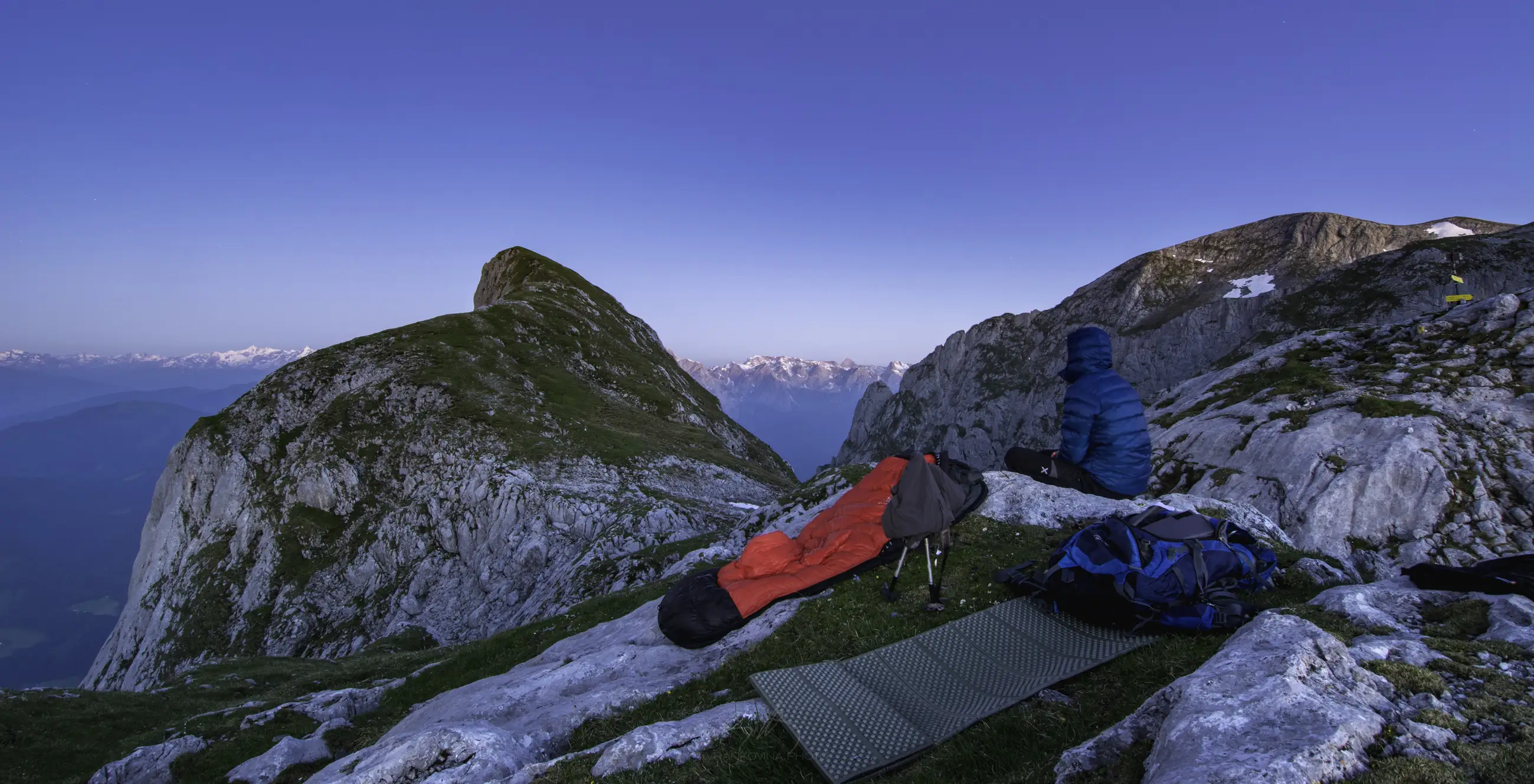 A person in a blue jacket sits on a rocky mountain slope at sunrise, beside a red sleeping bag and backpack, overlooking distant snow-capped peaks under a clear blue sky.