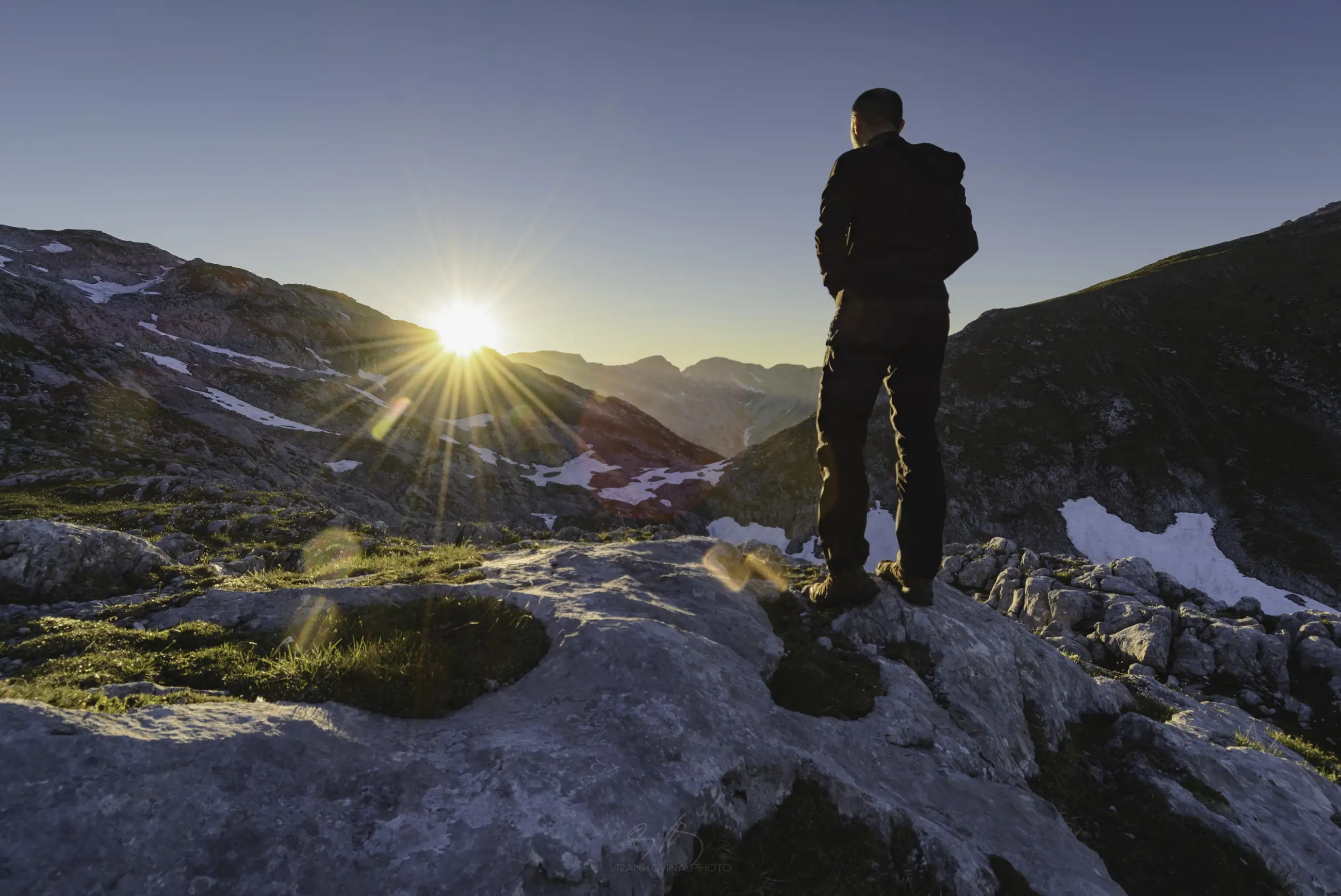 A person stands on a rocky mountain top at sunrise, overlooking a scenic landscape of distant peaks and patches of snow under a clear sky.