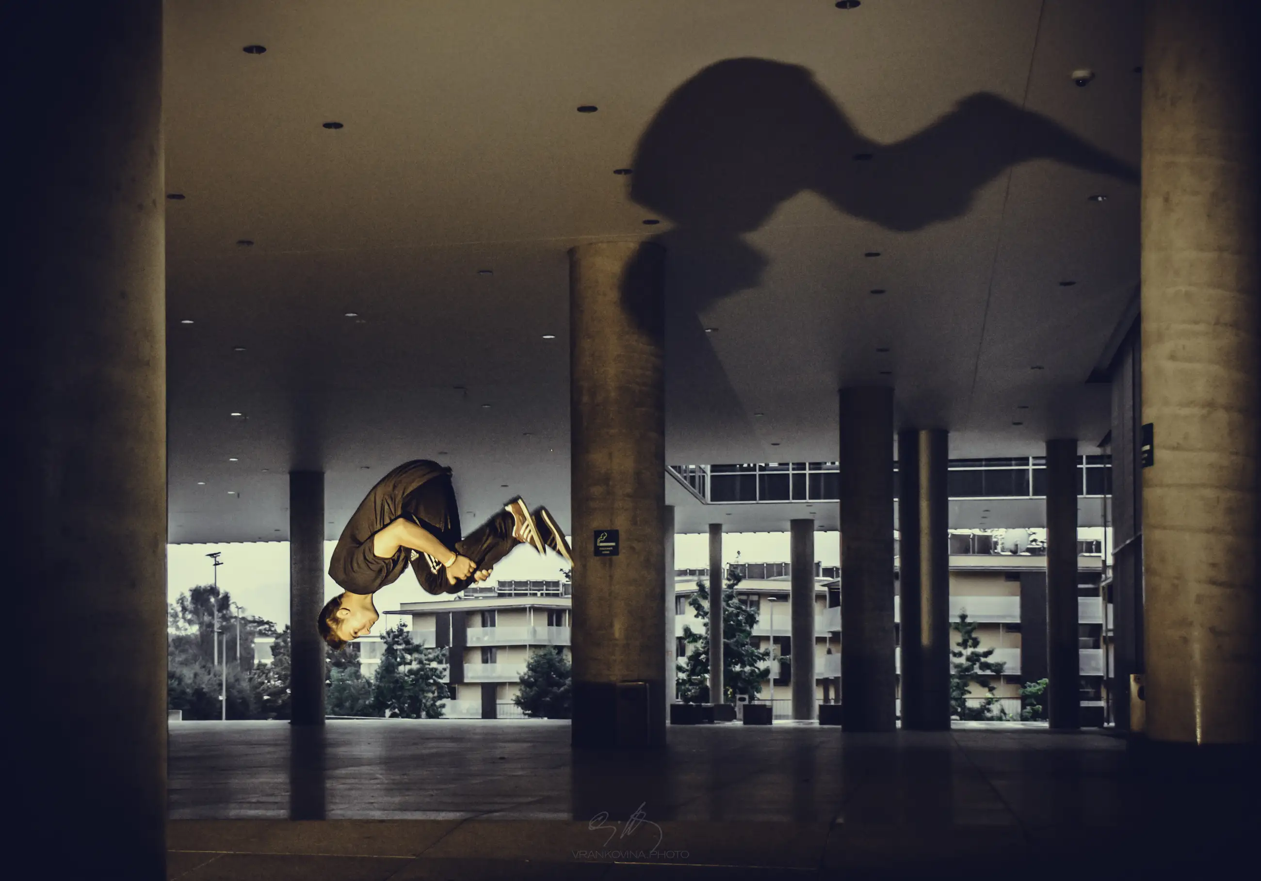 A person performs a backflip in midair under a covered outdoor area, casting a large, dramatic shadow on the ceiling. Numerous columns and buildings are visible in the background.