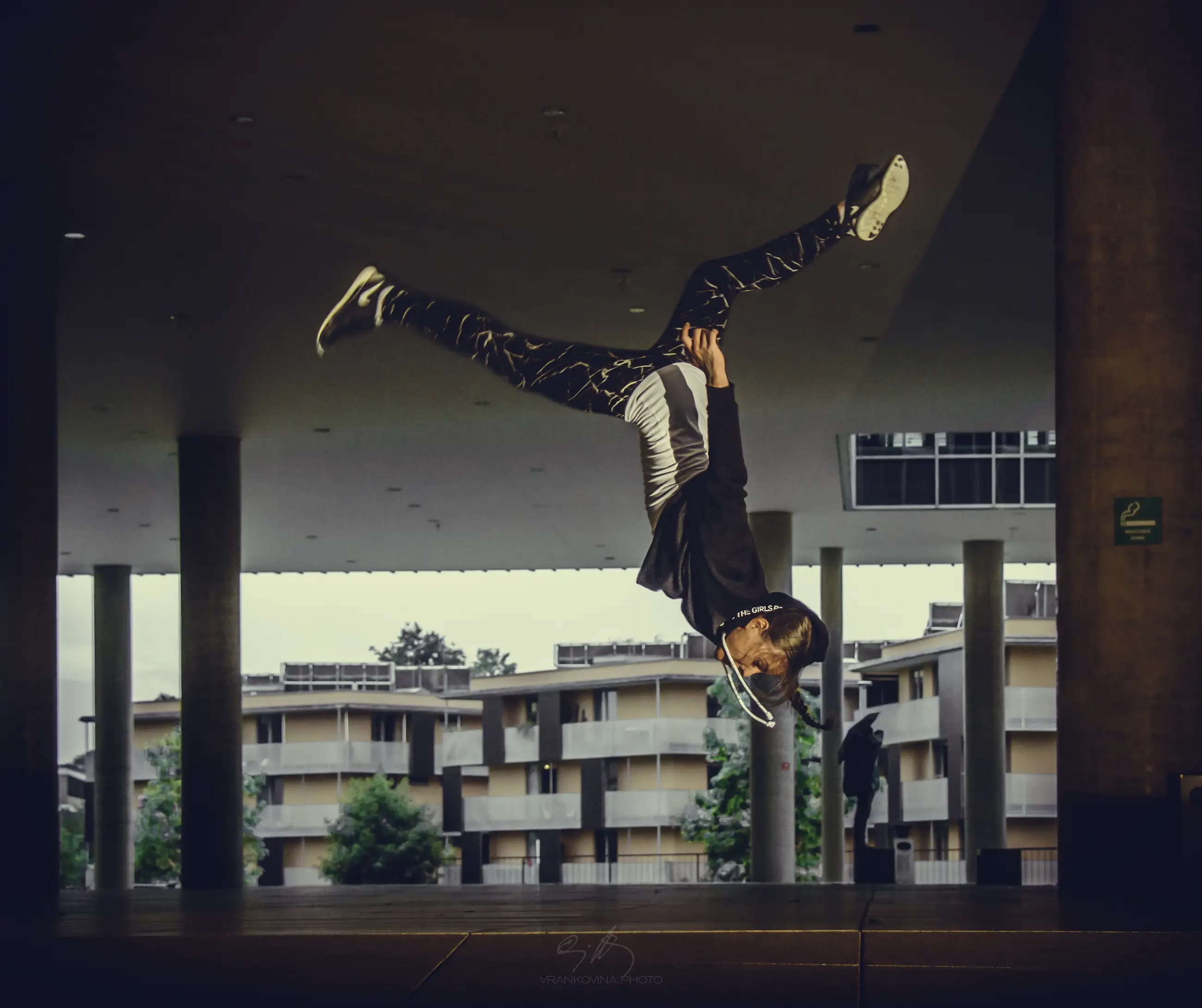 A person in athletic clothing performs a dramatic handstand flip indoors, with legs spread wide in midair. Modern buildings and columns are visible in the background.