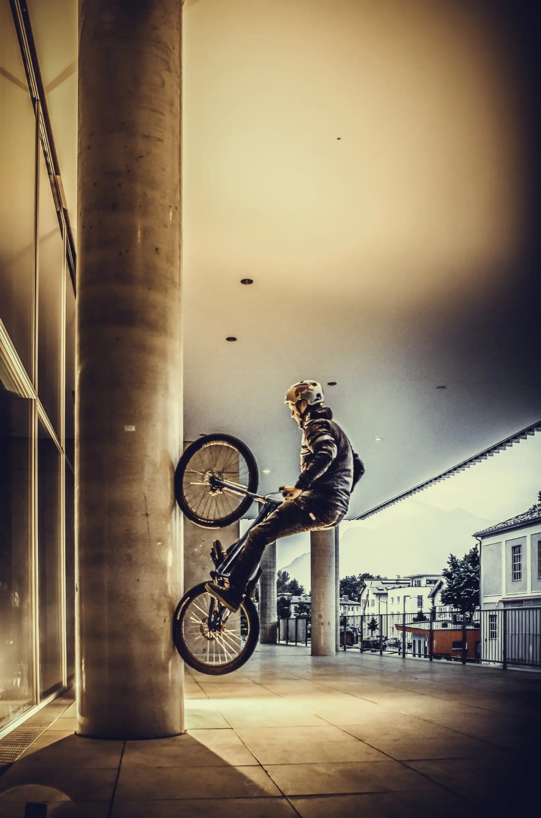 A person wearing a helmet performs a bike stunt, riding vertically up a concrete column in an urban area, with buildings and glass windows visible in the background.