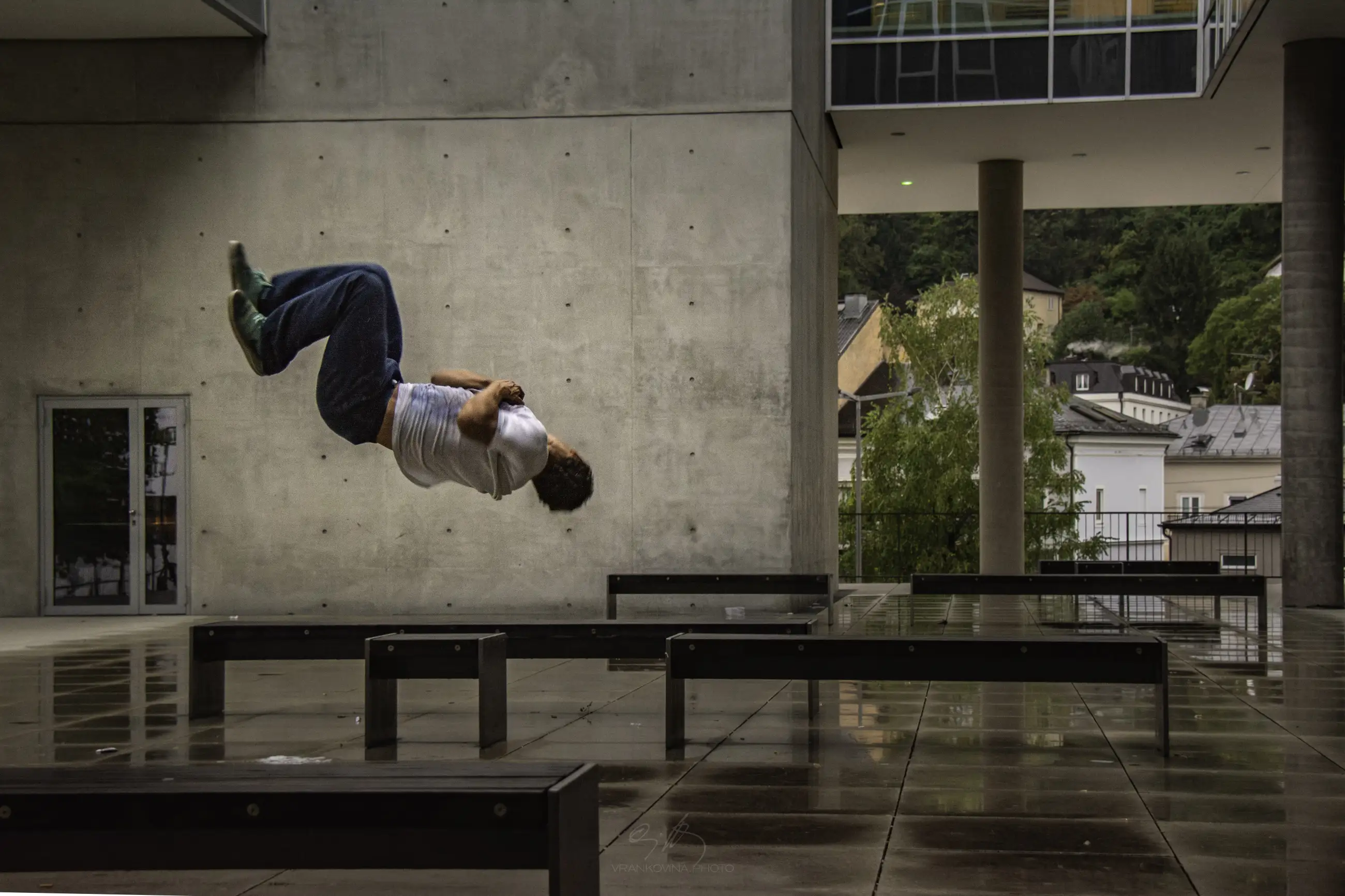 A person in a gray shirt and jeans performs a backflip over metal beams on a wet, outdoor urban plaza with modern concrete buildings and trees in the background.