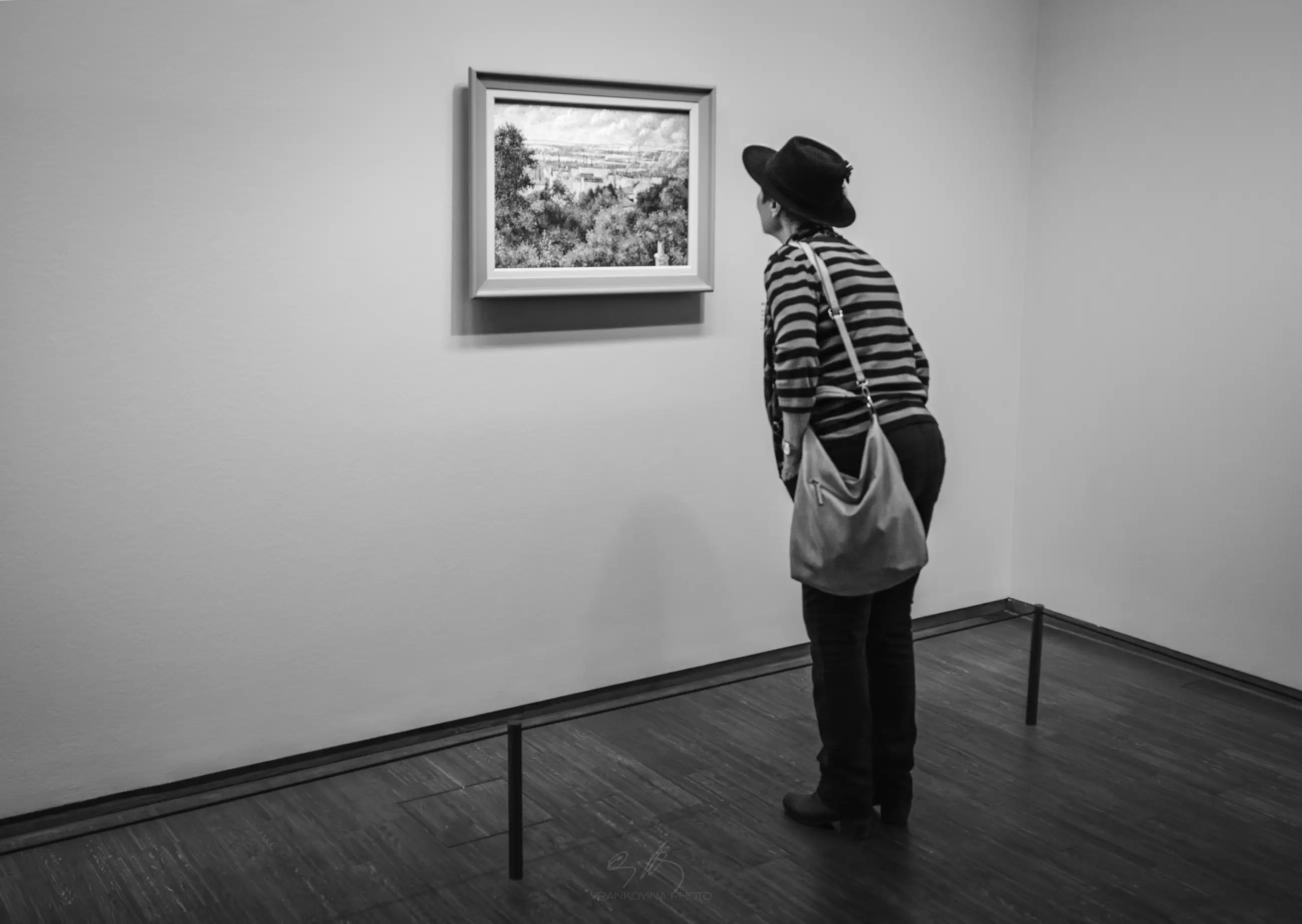 A person wearing a striped shirt, hat, and shoulder bag closely observes a framed landscape painting on a white gallery wall. The scene is in black and white.