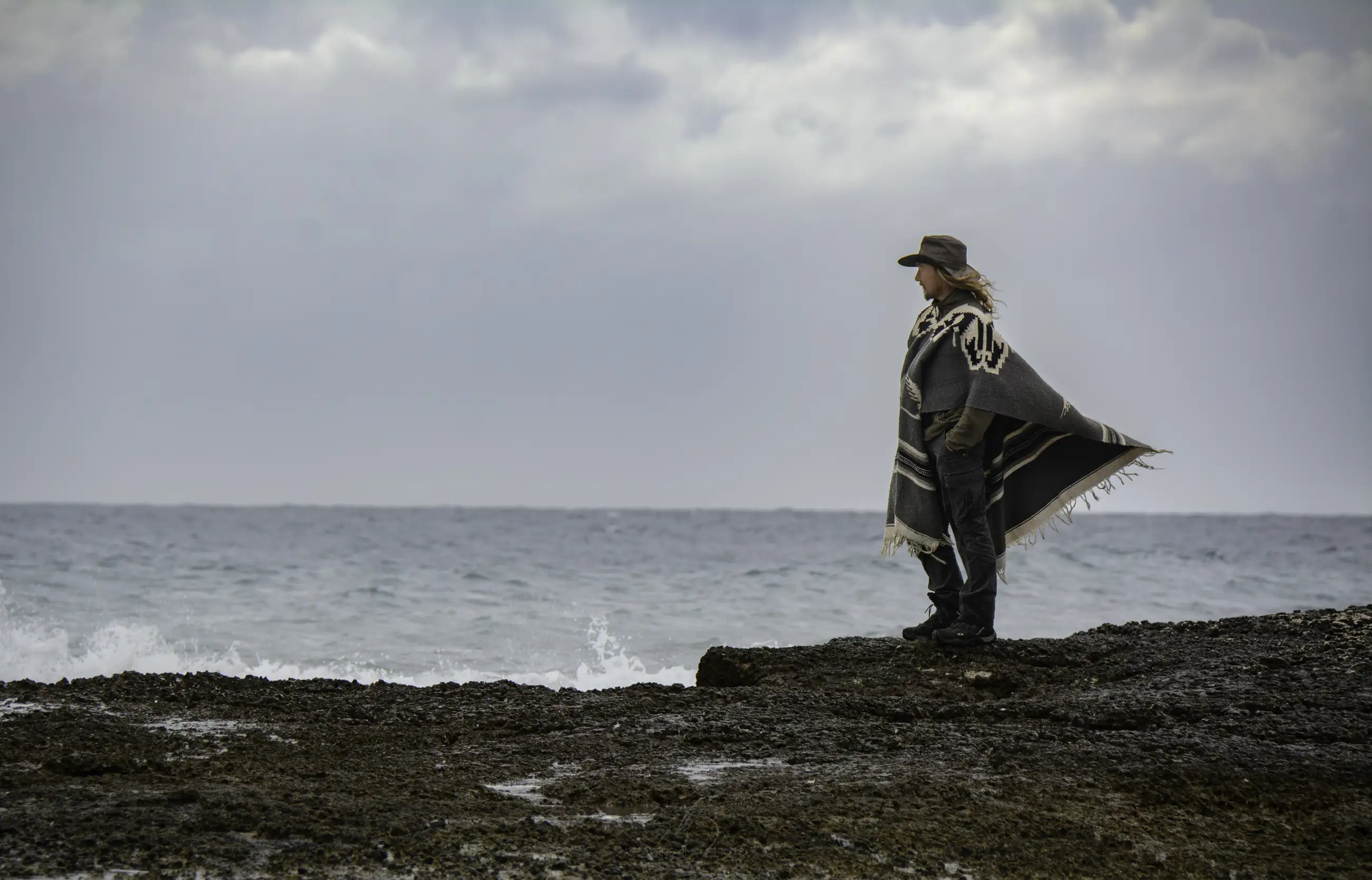 A person wearing a hat and a poncho stands on a rocky shore, looking out at the ocean under a cloudy sky. Waves hit the rocks nearby.