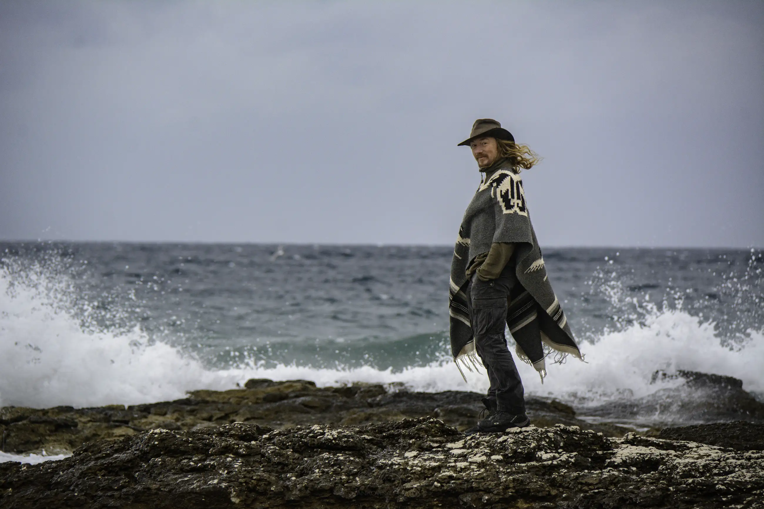 A person wearing a hat and a patterned poncho stands on rocky shore with ocean waves crashing behind them under a cloudy sky.