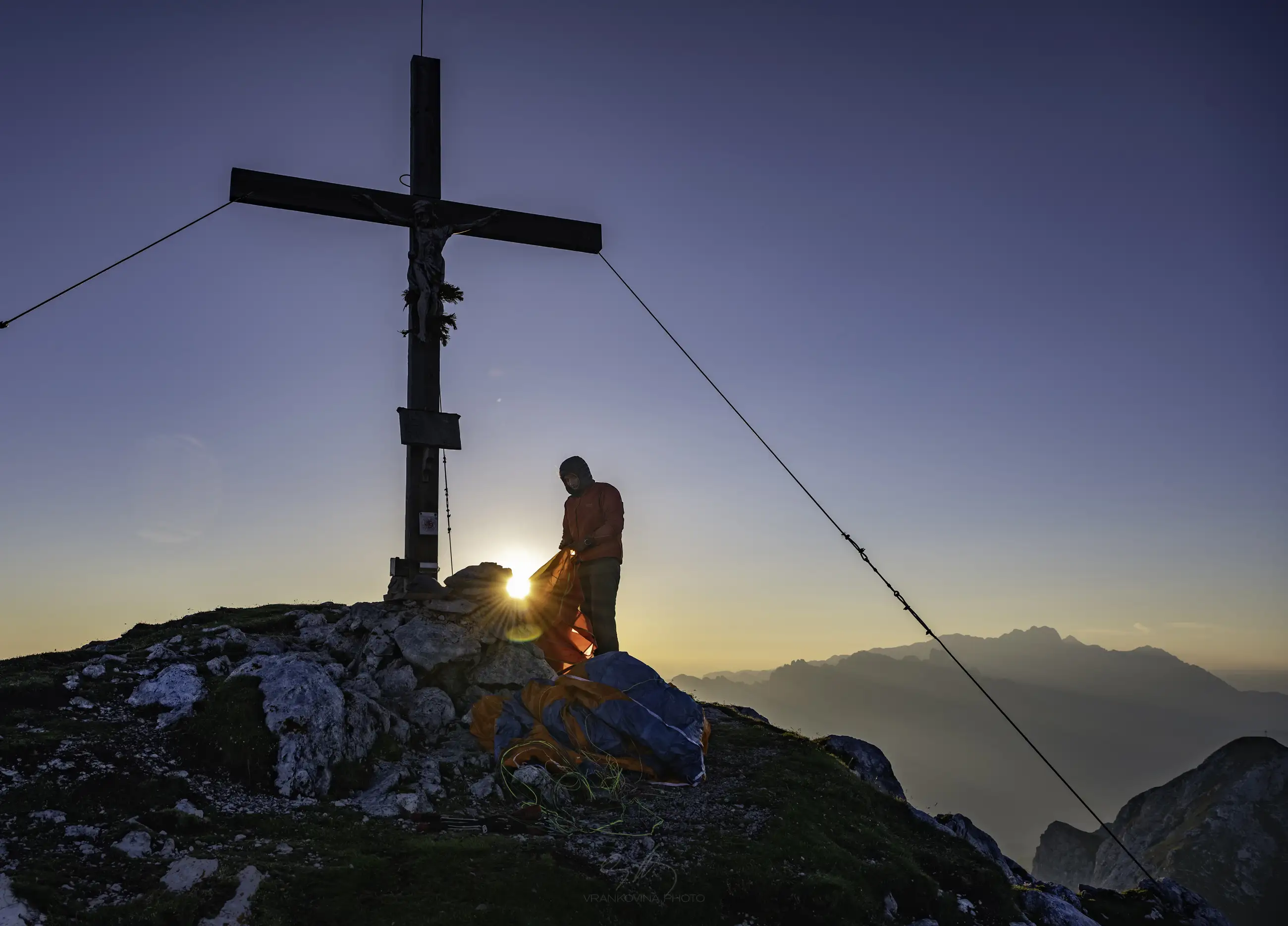 A person stands beside a large wooden cross at a mountain summit during sunrise, with camping gear on the ground and distant peaks visible under a clear sky.