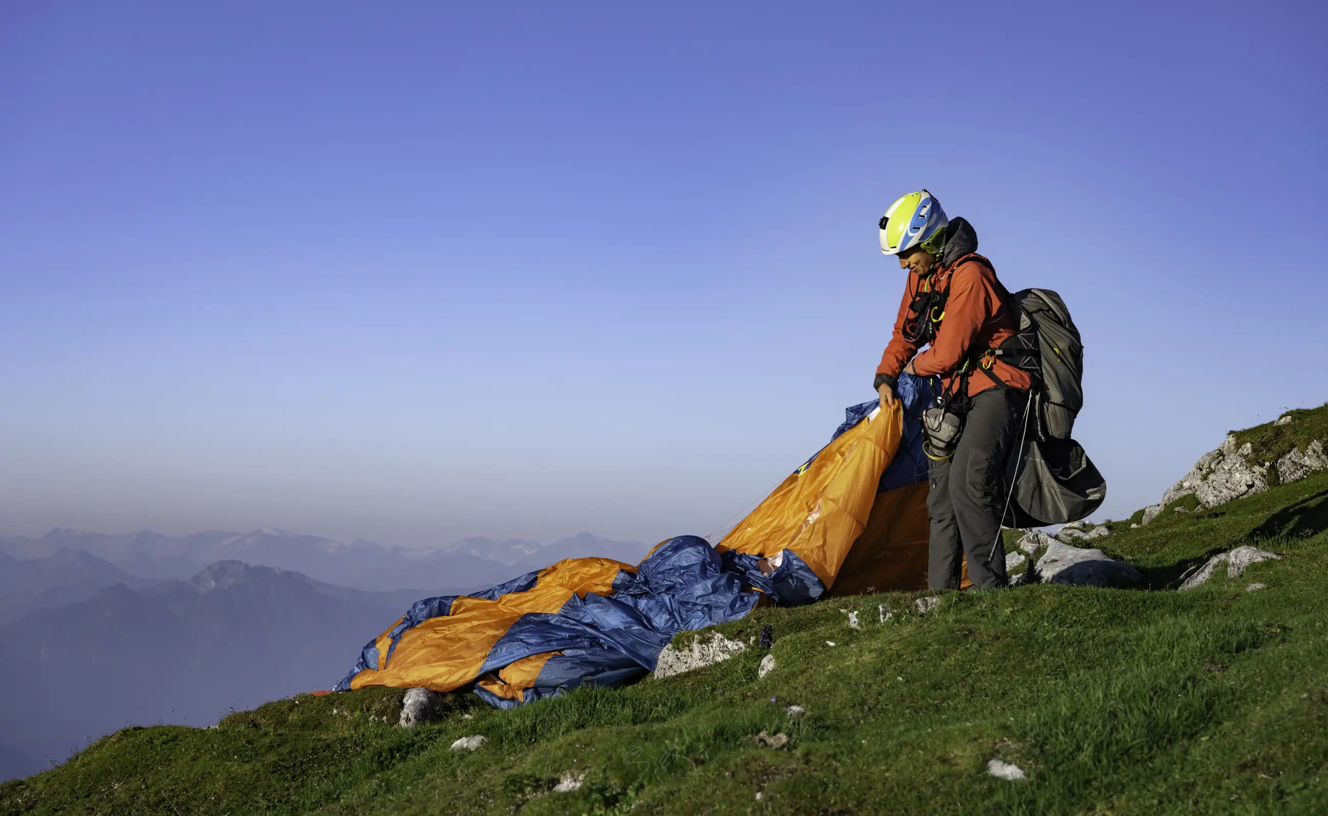A person in outdoor gear and helmet prepares an orange and blue paraglider on a grassy hillside, with mountains and a clear blue sky in the background.