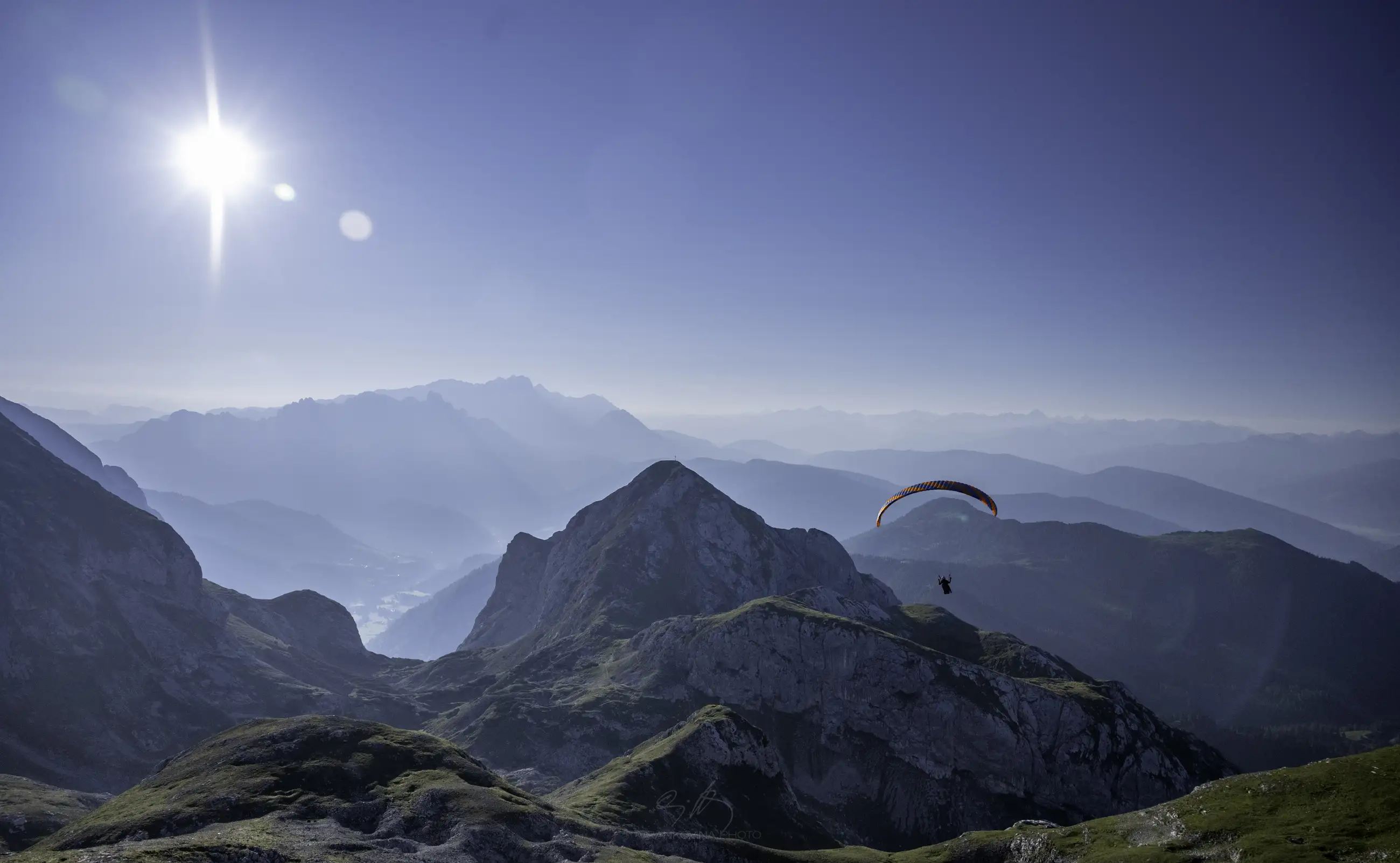 A paraglider soars above rugged mountain peaks under a clear blue sky, with the sun shining brightly and layers of distant mountains fading into the horizon.