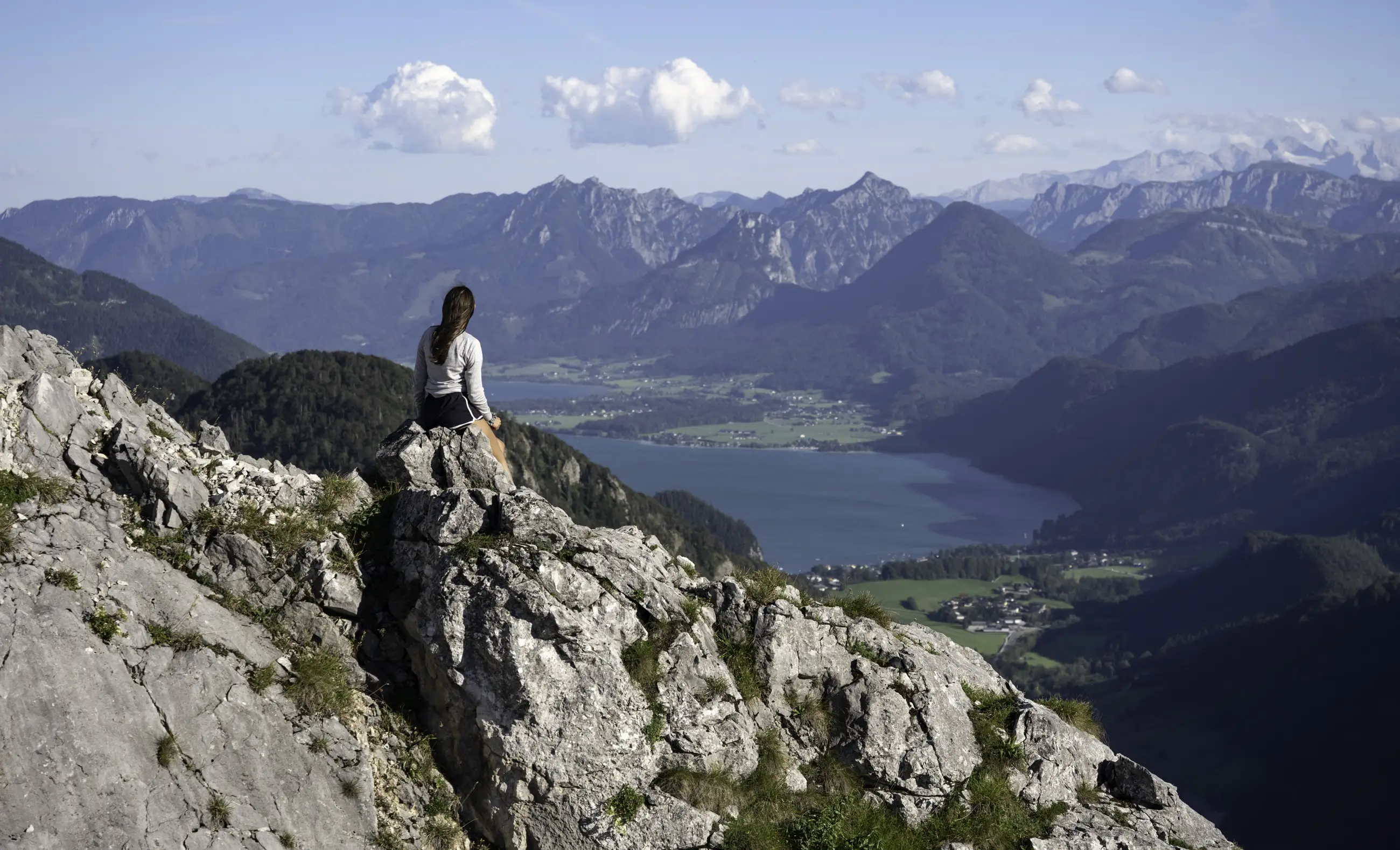 A woman sits on a rocky outcrop overlooking a scenic valley with a lake, green fields, and distant mountains under a blue sky with scattered clouds.