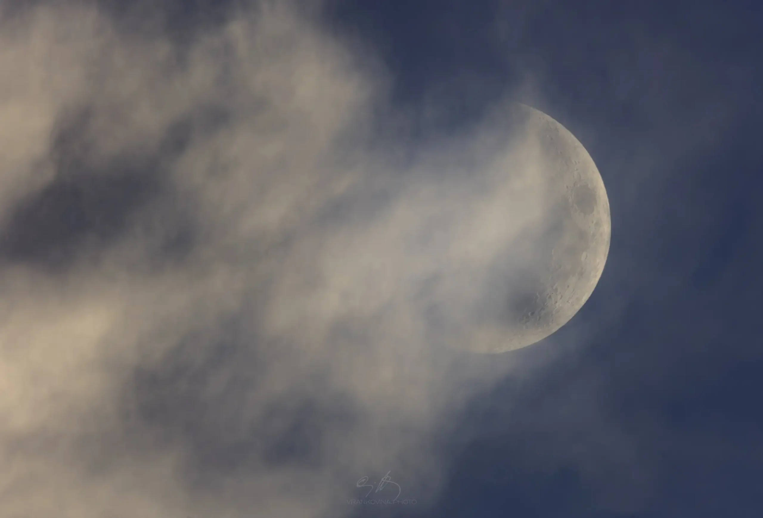 A waxing gibbous moon partially obscured by thin, wispy clouds in a blue sky.