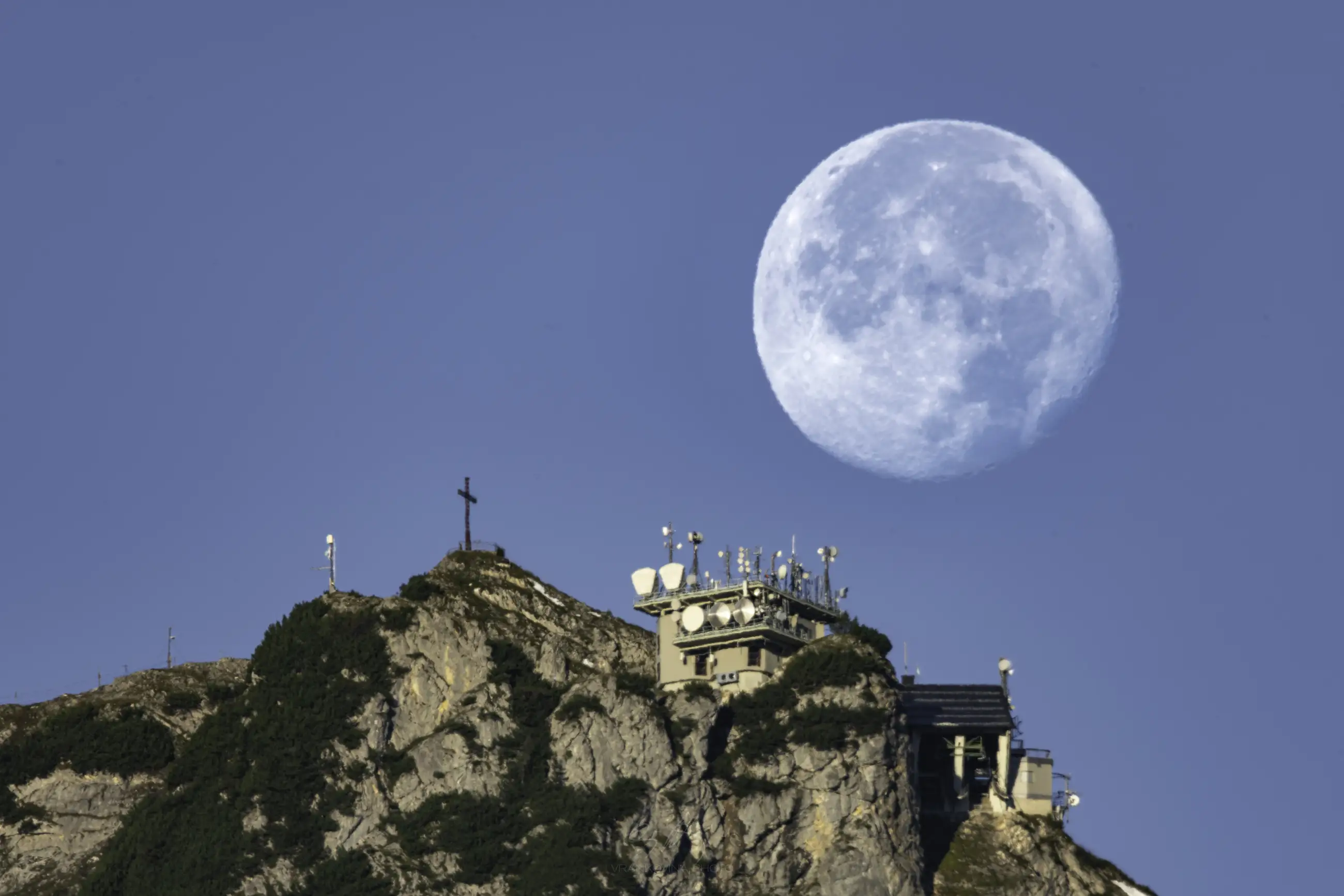 A large, bright full moon rises behind a rocky mountain peak with a building, antennas, and a cross on top, all set against a clear blue sky.