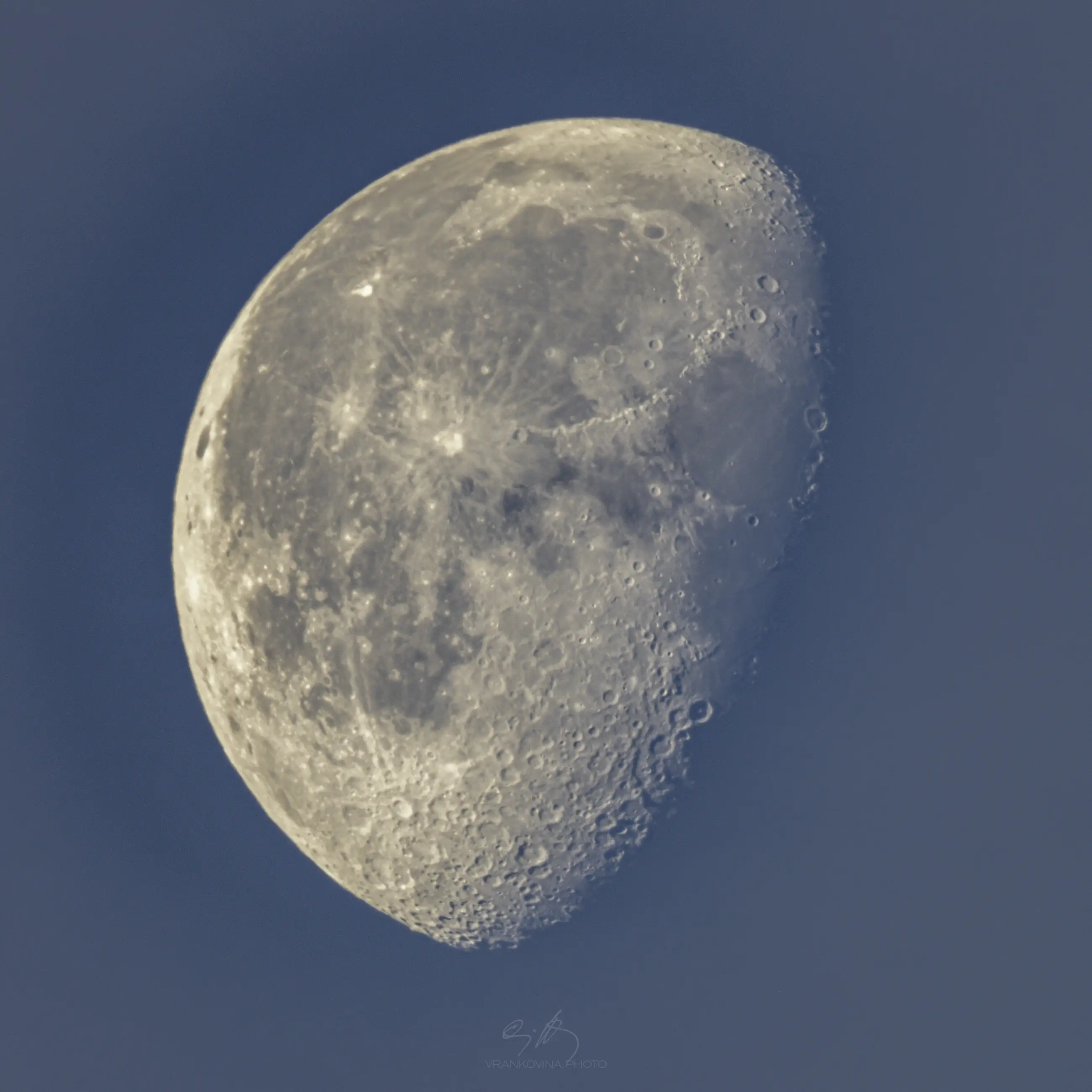 A close-up photo of a waxing gibbous moon against a blue sky, showing detailed craters and surface textures.