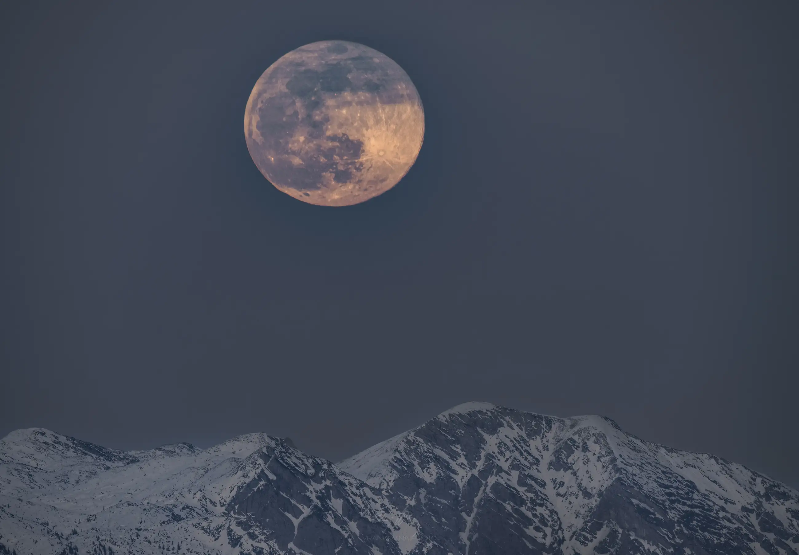 A large, reddish full moon rises above snow-capped mountain peaks under a clear, dusky sky.
