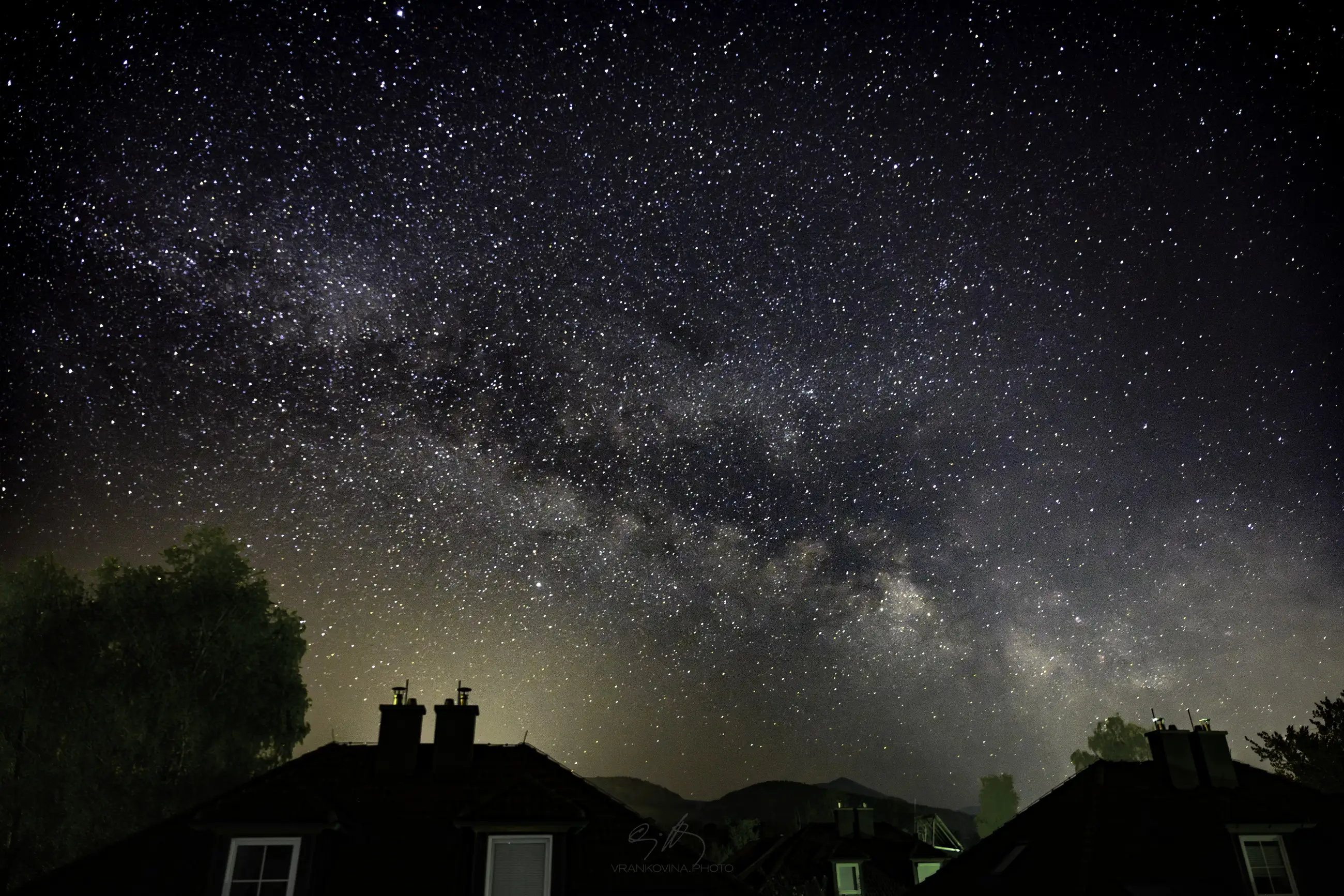A night sky filled with stars and the Milky Way stretching above dark silhouettes of rooftops, trees, and distant hills, with a faint glow on the horizon.