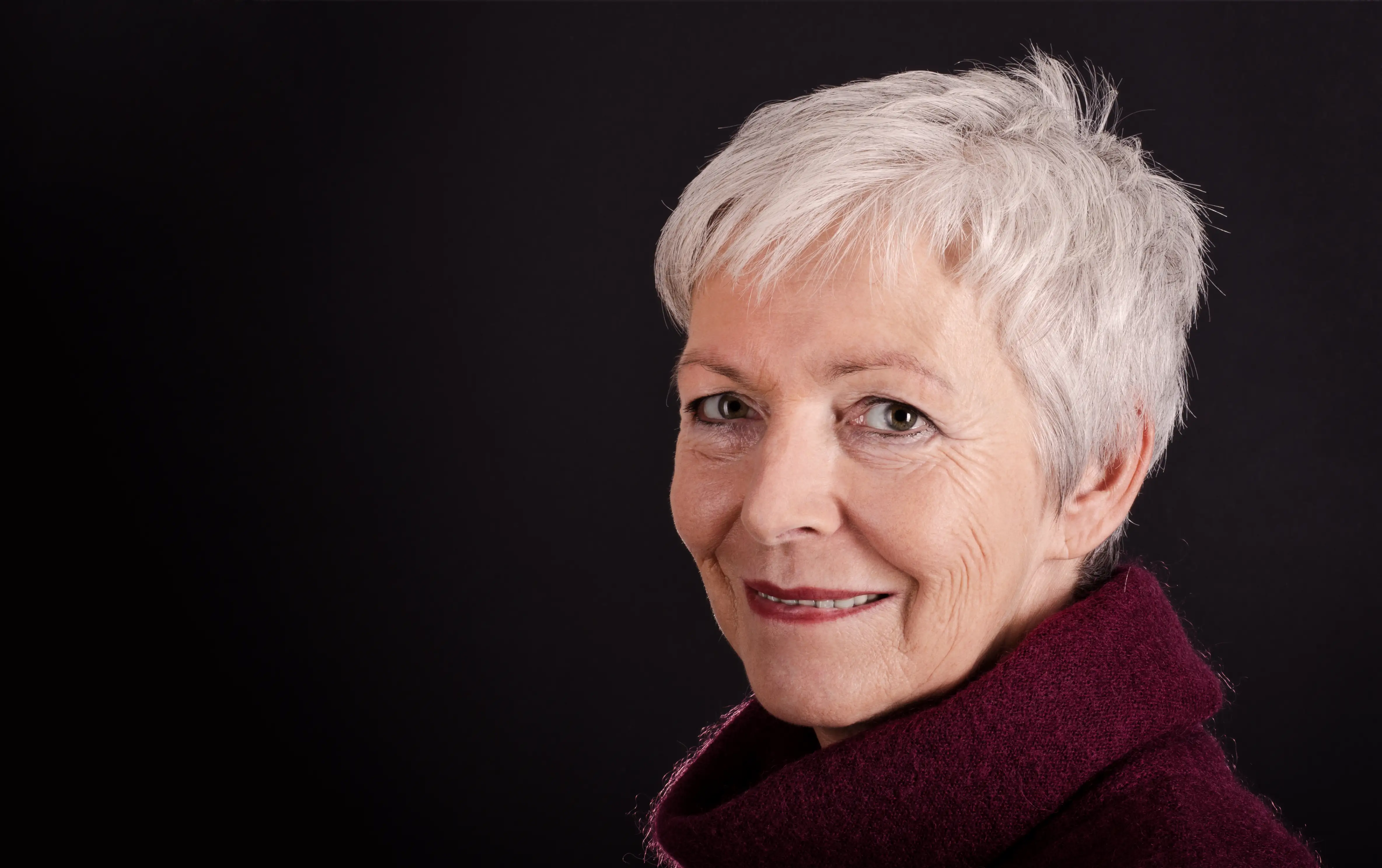 An older woman with short white hair and light eyes, wearing a burgundy sweater, smiles gently at the camera against a plain black background.